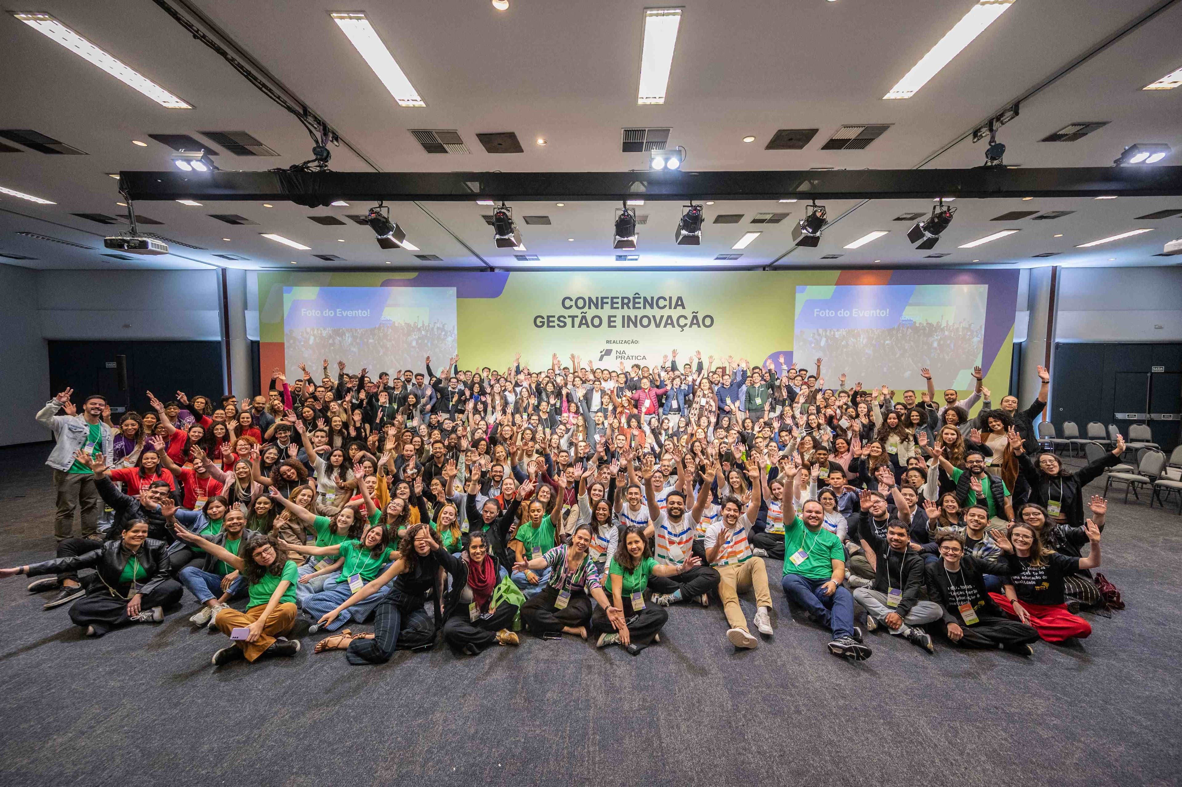 Foto mostra pessoas participantes da Conferência Na Prática Gestão e Inovação com mãos para o alto e reunidas diante do palco do evento