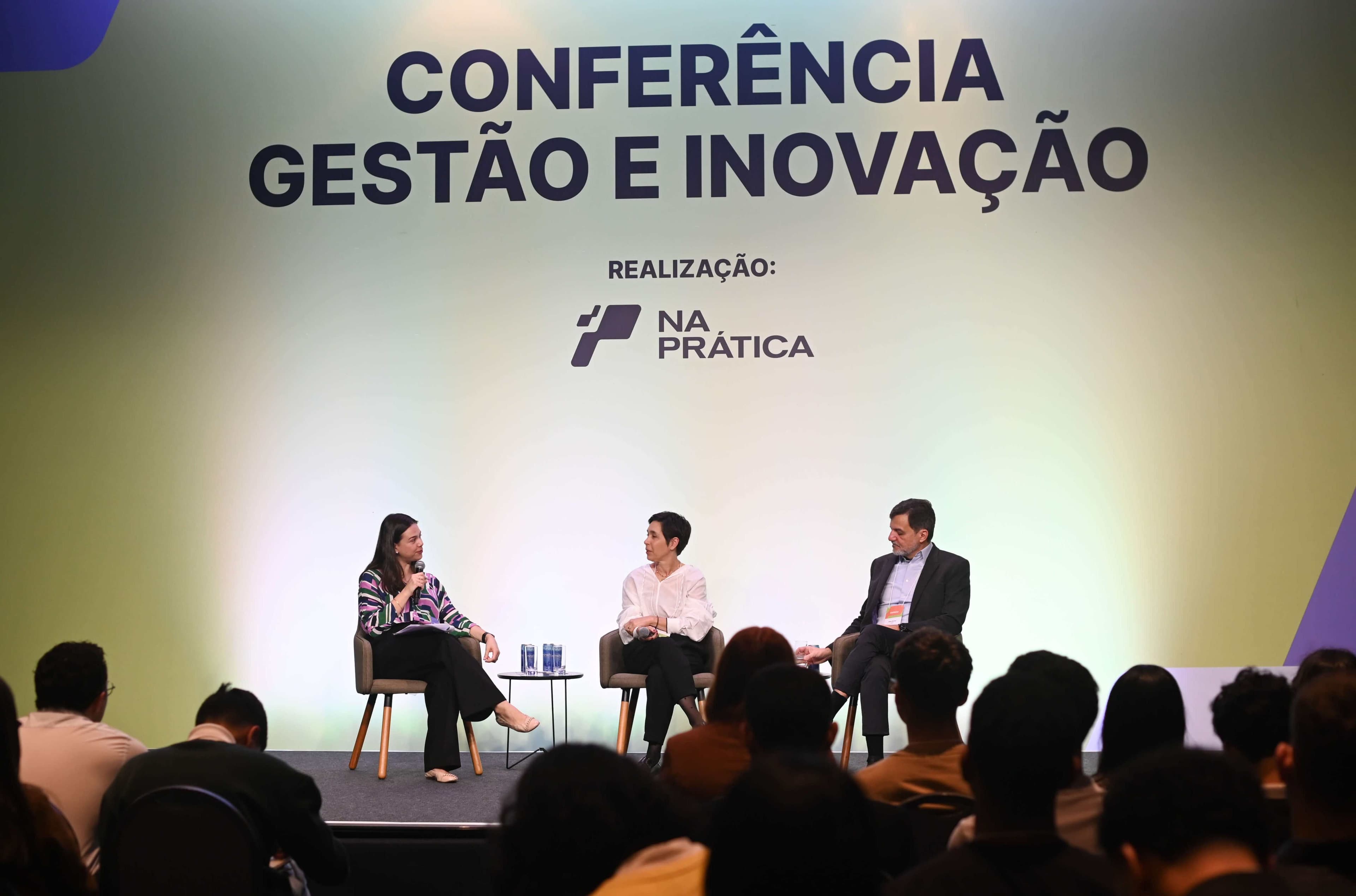 Foto mostra trio de painelistas no palco da Conferência Gestão e Inovação do Na Prática. Anamaíra Spaggiari é uma mulher branca, de cabelo castanho e longo, que veste calças pretas e blusa social colorida. Lorice Scalise, sentada em cadeira ao lado de Anamaíra, é uma mulher magra, de cabelos curtos e pretos, que veste camisa social branca e calça social preta, e Líbano Barroso, sentado ao lado de Scalise, é um homem branco de cabelos curtos que veste terno cinza, sem gravata.