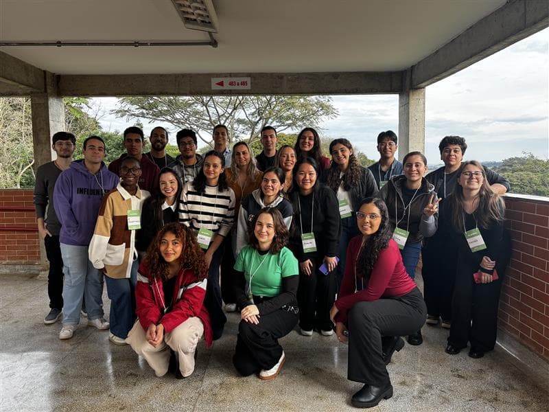Foto em grupo de aproximadamente 25 pessoas, entre jovens e adultos, posando para a câmera em um espaço coberto com estrutura de concreto e paredes de tijolo aparente. O local parece ser um mirante ou área de descanso, com vista para uma paisagem verde e montanhosa ao fundo sob um céu nublado. Os participantes vestem roupas casuais em cores variadas, alguns usando crachás com fita verde, sugerindo tratar-se de um evento acadêmico ou profissional. O grupo está distribuído em três fileiras - alguns em pé na parte de trás, outros sentados no meio e três pessoas agachadas na frente. Todos demonstram expressões alegres e descontraídas, indicando um momento de confraternização ou encerramento de atividade.