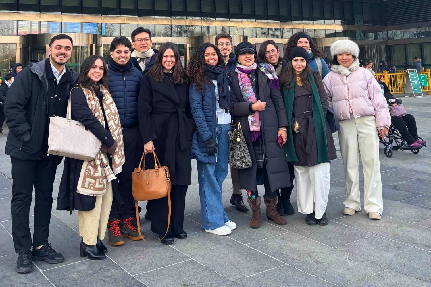 Foto com grupo de estudantes na frente de hospital na china. Nome do hospital está em mandarim, com letras vermelhas, em um edifício cinza. O grupo está agasalhado.