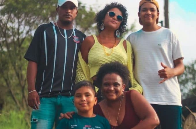 Uma família posa para uma foto. Na parte superior, um homem jovem com a camisa preta do Coritnhians, uma mulher com vestido verde e um garoto com uma camisa branca. Na parte inferior, Talísia abraça seu filho mais novo, uma criança de aproxidamente 9 anos.