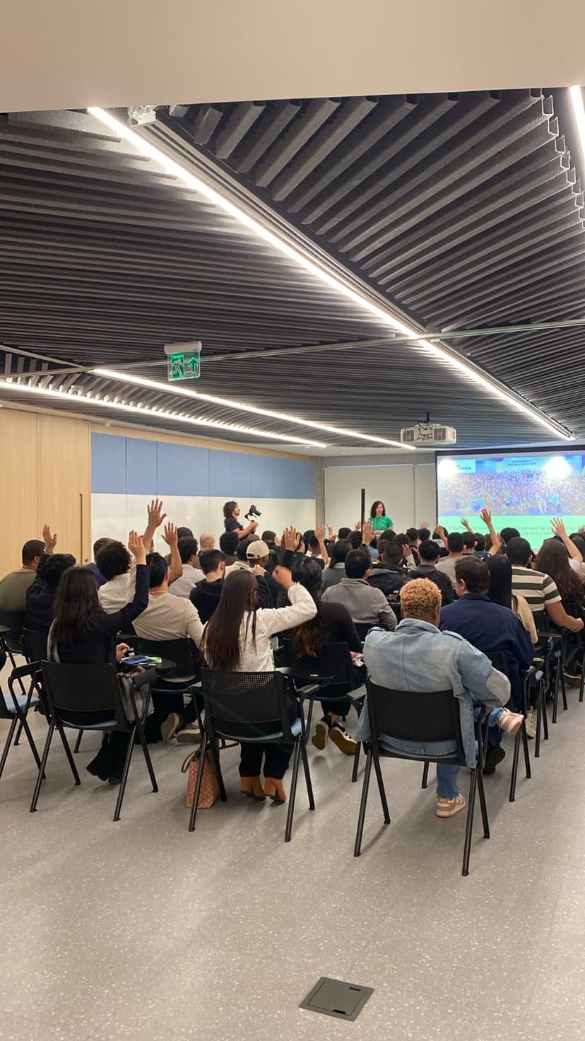 Foto mostra alunos sentados em sala de aula diante de facilitadora. Parte deles tem os braços levantados.