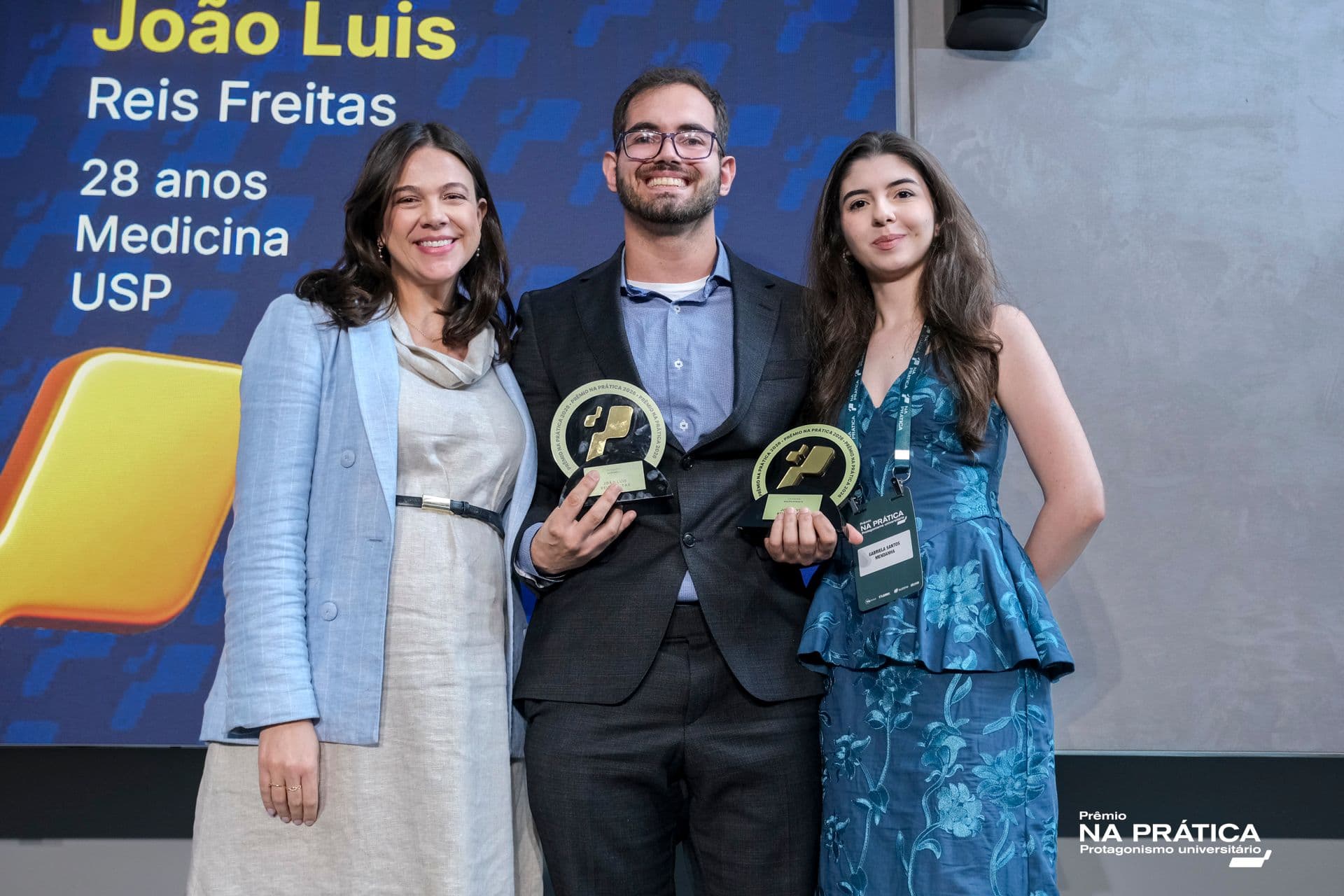 Jovem posa para foto ao lado de duas mulheres. Ele segura dois troféus redondos
