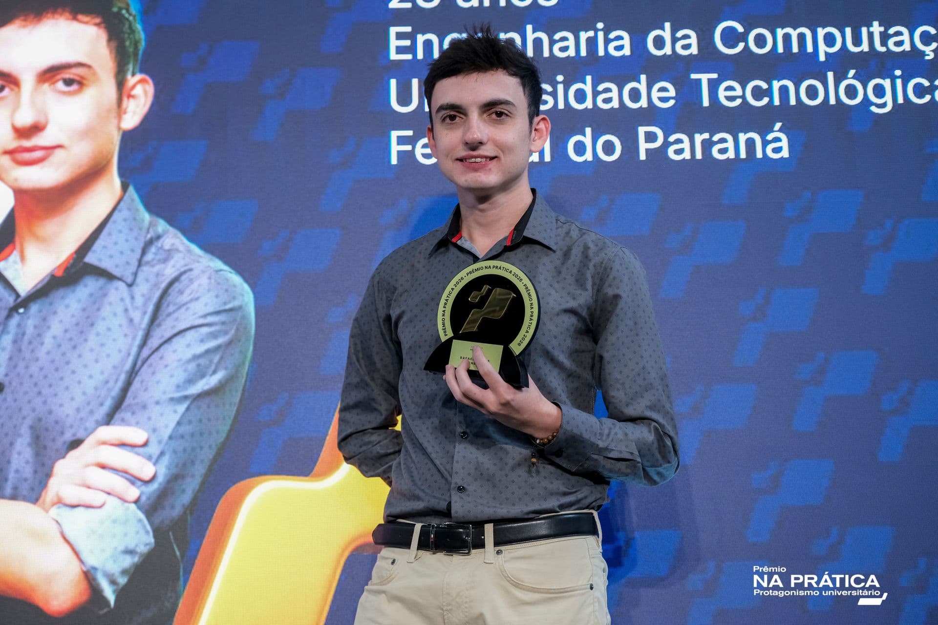 Jovem de camisa cinza segura troféu em frente a painel com própria foto dele