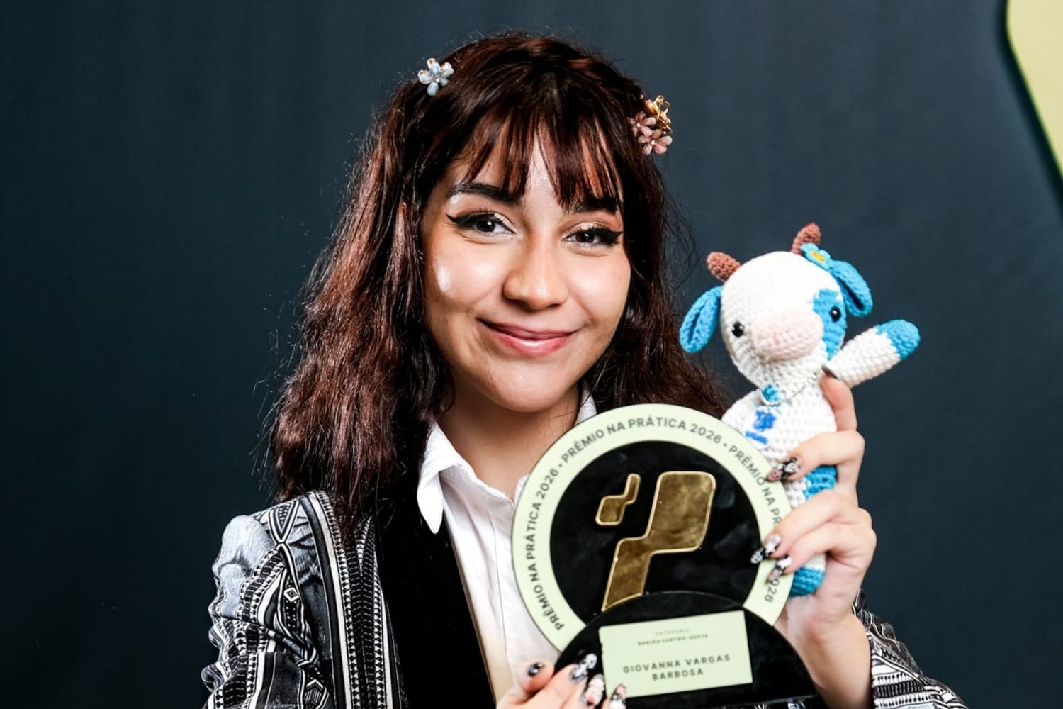 Jovem branca, vestida de roupa social, segurando um troféu redondo e um vaquinha azul de pelúcia