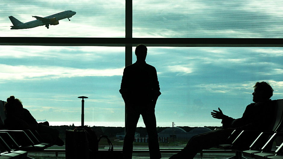 Homens aguardam voo em sala de espera em aeroporto. Pela janela, assistem avião decolar