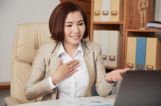 Young Asian woman in office wearing headphones and having online call via laptop sitting at table