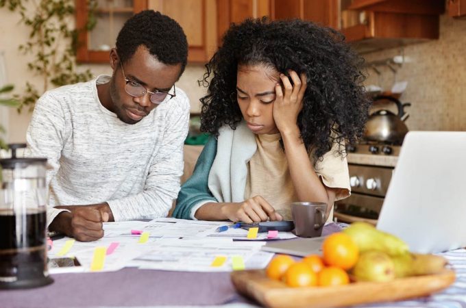 Financial problem and economic crisis concept. AngryAfrican male in glasses having stressed and puzzled expression, thinking over numerous debts, his unhappy wife sitting next to him and crying