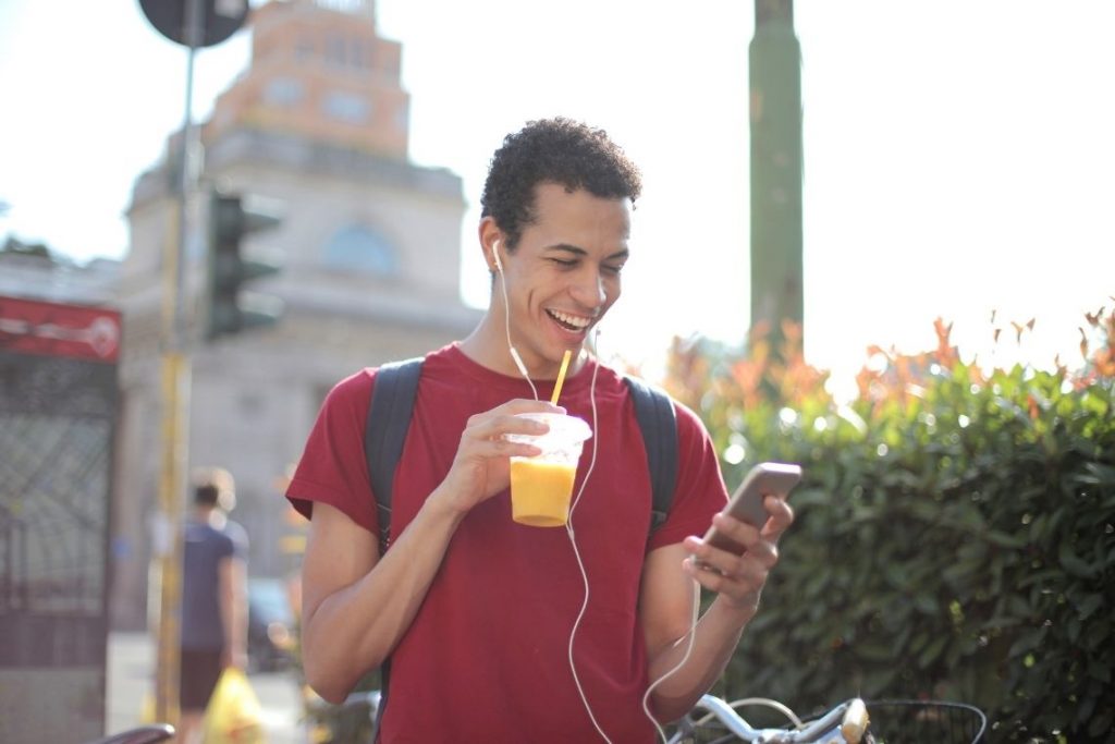 jovem homem de fone e tomando suco sorri enquanto olha para celular