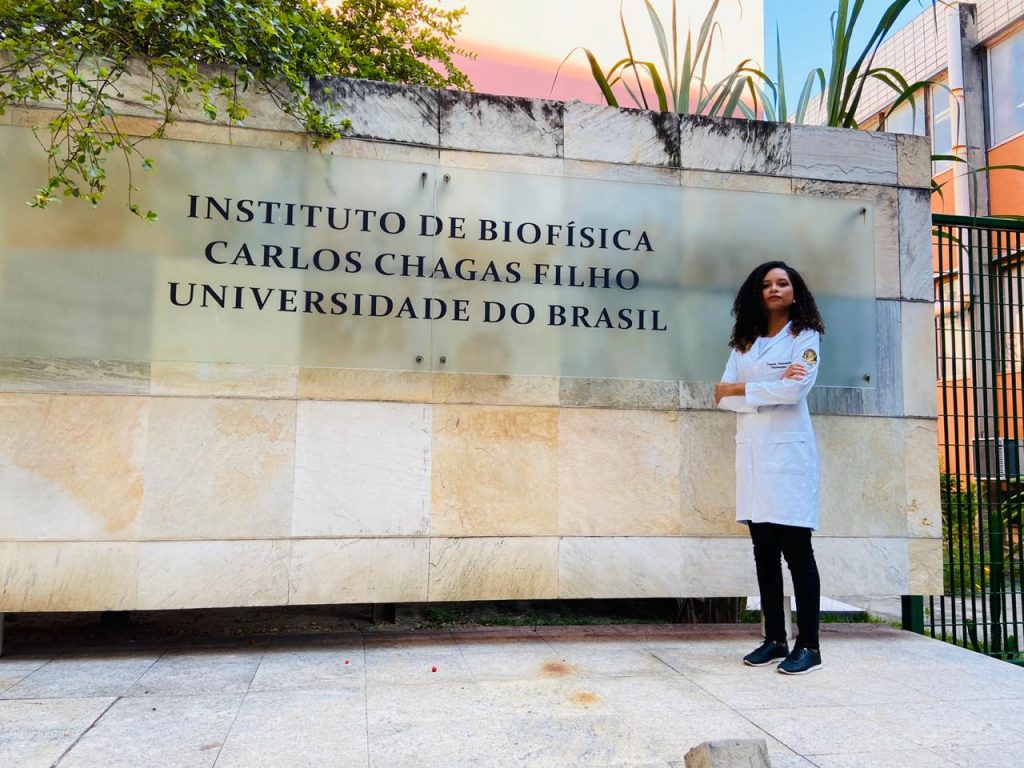 A jovem Dayene Caldeira, de 28 anos, posa de jaleco branco ao lado de um letreiro que diz: "Instituto de Biofísica Carlos Chagas Filhos. Universidade do Brasil". Ela é uma mulher negra, de cabelo crespo.