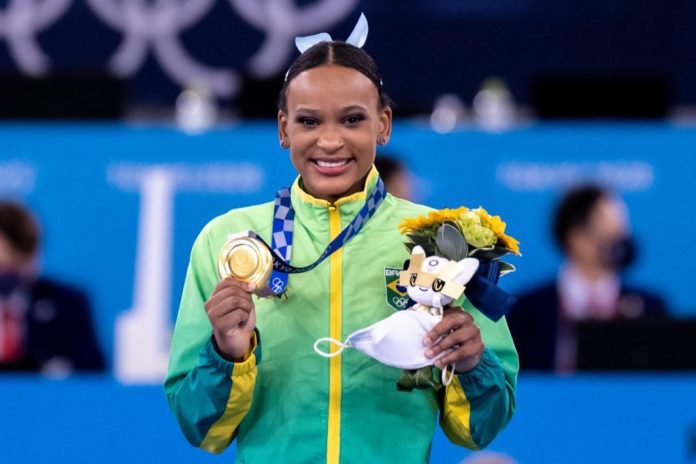 Rebeca Andrade é uma mulher jovem, negra, que usa o uniforme do Brasil durante a cerimônia de premiação das Olímpiadas de Tóquio. Ela exibe uma medalha de ouro e carrega um buquê de flores no centro da imagem, em destaque.