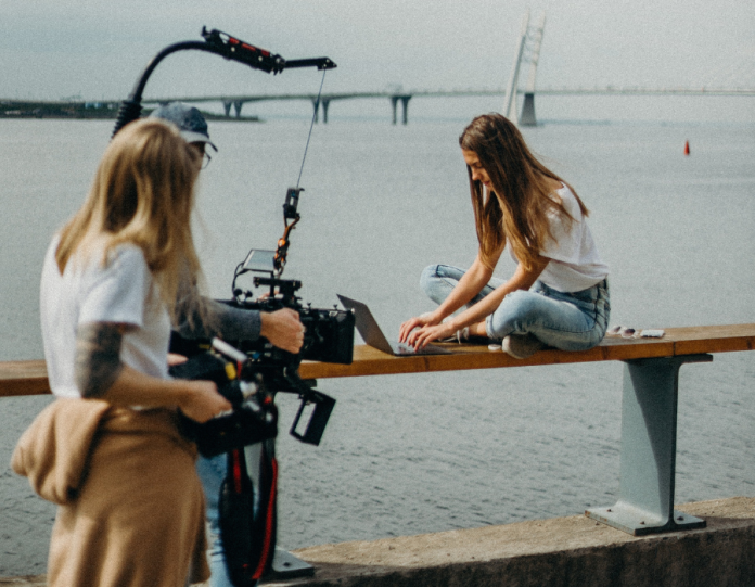 Uma mulher com uma câmera filma uma jovem em um banco à beira da praia. Um homem de boné dirige a cena.