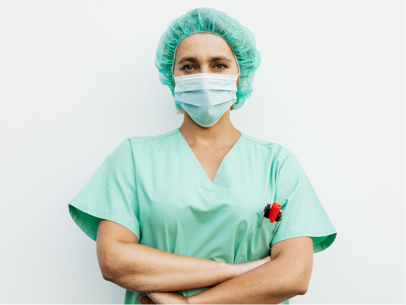 Imagem mostra profissional da enfermagem vestida com seu uniforme típico, máscara e touca. No bolso da frente da blusa, ela carrega canetas.