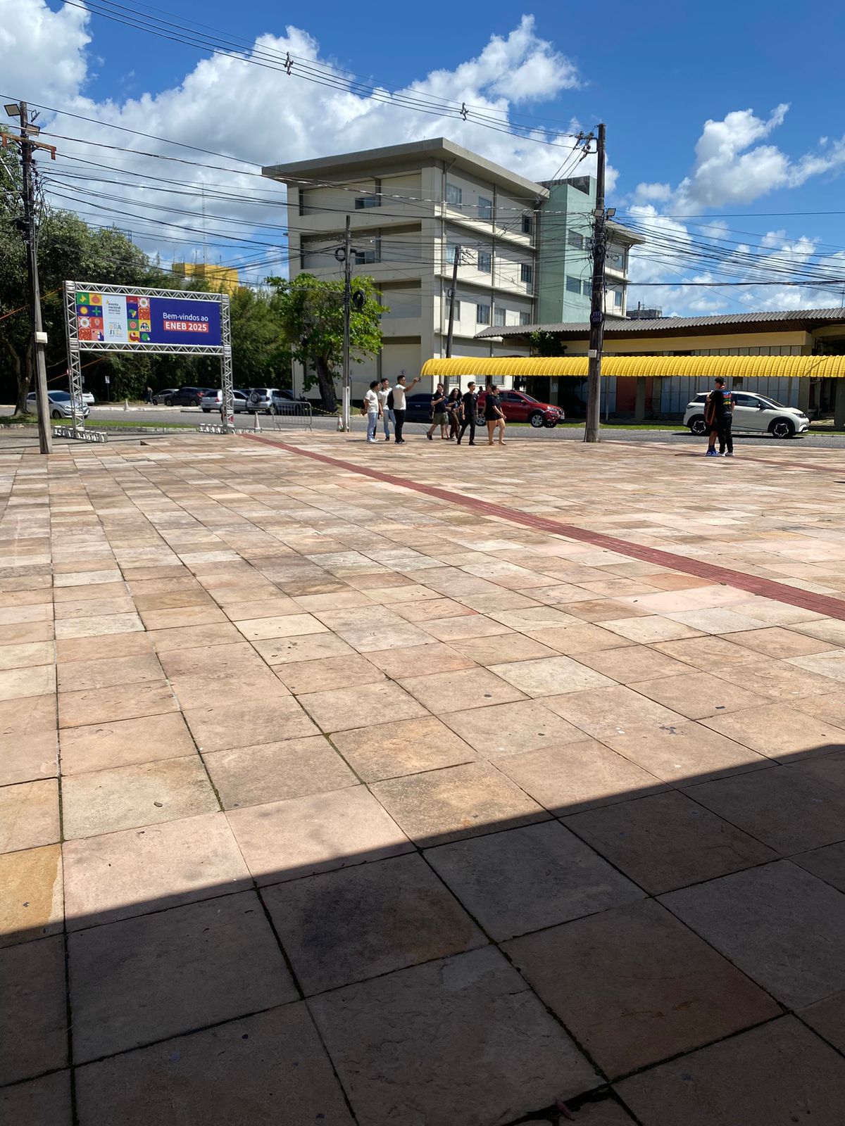 A imagem mostra uma praça urbana pavimentada com pedras em tons bege e marrom, apresentando um padrão geométrico com faixas vermelhas. No centro da praça há algumas pessoas caminhando e se reunindo. Ao fundo, observa-se um portal de estrutura metálica com uma placa colorida que menciona "ENEM 2025". A cena inclui edifícios residenciais de múltiplos andares, uma construção comercial térrea com toldo amarelo, postes de energia elétrica com vários fios, árvores e veículos estacionados. O céu está parcialmente nublado com nuvens brancas espalhadas sobre um fundo azul, criando uma atmosfera típica de um dia ensolarado em área urbana brasileira.